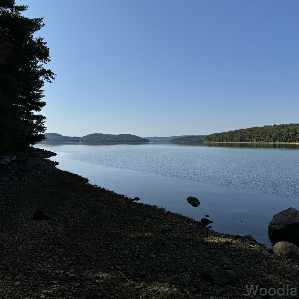 Glass-smooth water along the shoreline of Quabbin Reservoir, contrasting with rippled water farther out, with islands and hills in the distance under a clear blue sky
