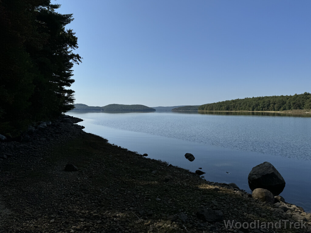 Glass-smooth water along the shoreline of Quabbin Reservoir, contrasting with rippled water farther out, with islands and hills in the distance under a clear blue sky