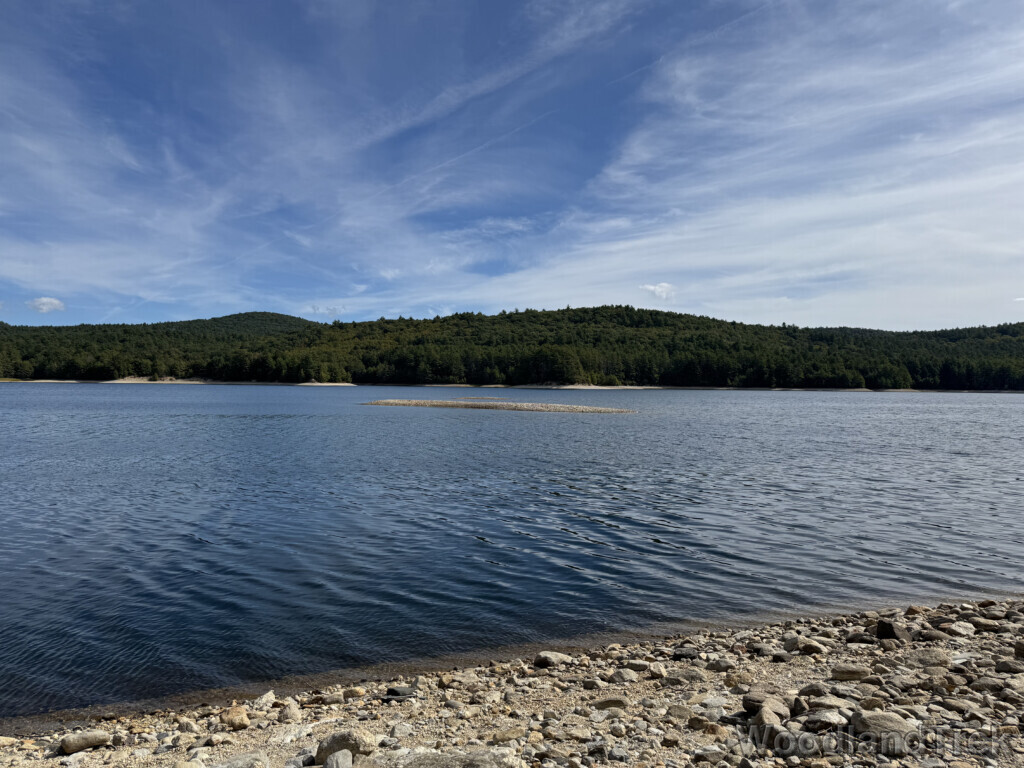Reservoir with calm water, forested shoreline, and hills in the distance under wispy cloud
